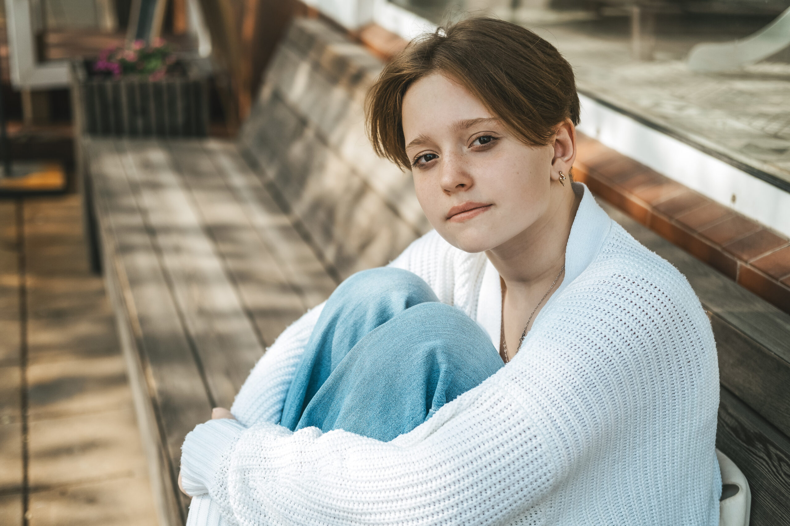 A teenage girl sits on a bench on the street, hugging her knees. Teen